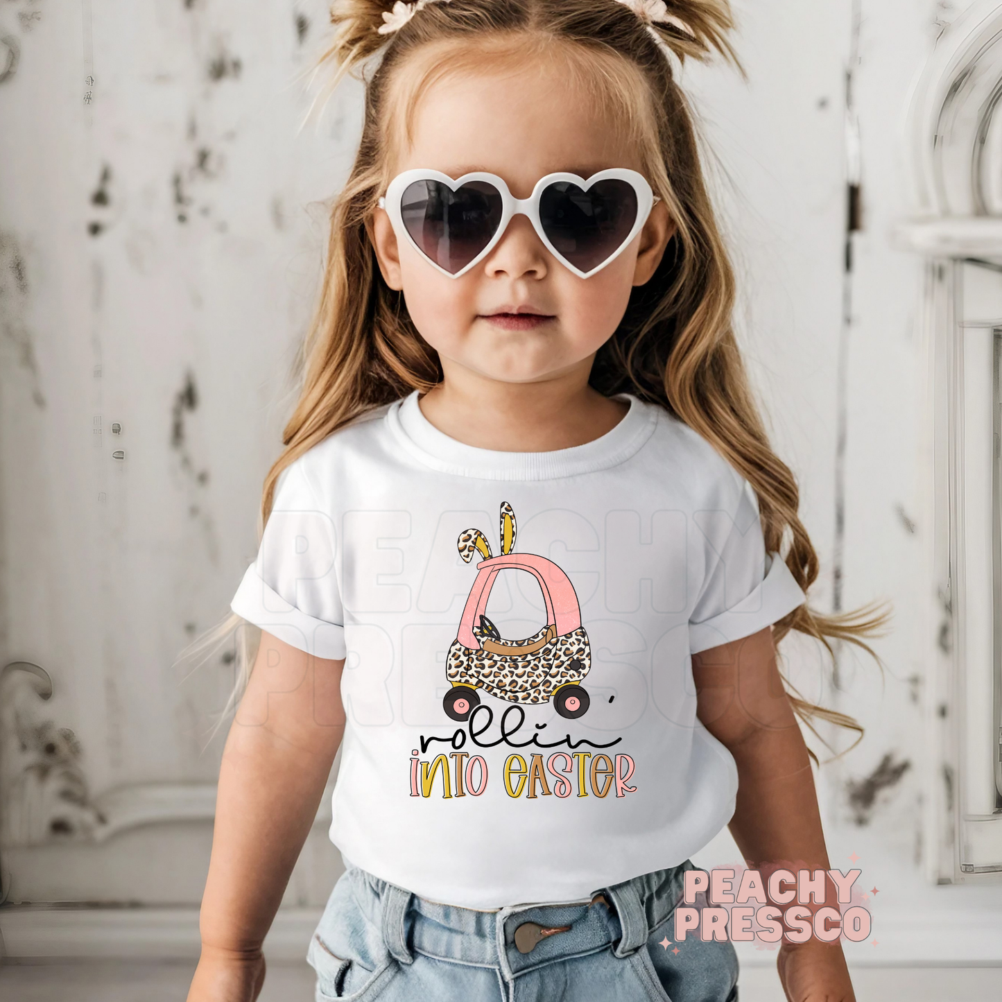 Toddler girl wearing a white Rollin Into Easter shirt featuring a pink bunny car with leopard print and Easter bunny ears.