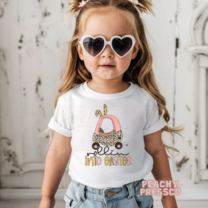 Toddler girl wearing a white Rollin Into Easter shirt featuring a pink bunny car with leopard print and Easter bunny ears.