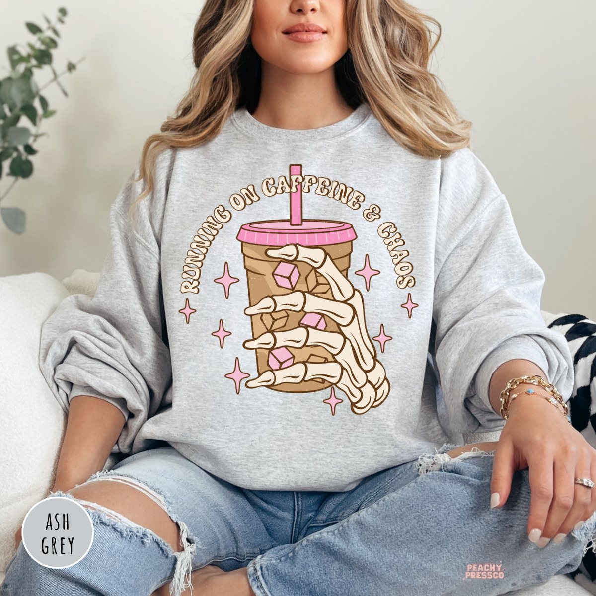 Woman wearing an ash grey crewneck sweatshirt featuring a skeleton hand iced coffee graphic with pink accents.