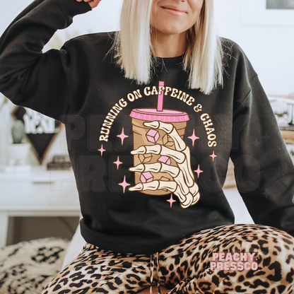 Woman wearing a black crewneck sweatshirt with a skeleton hand iced coffee graphic and retro caffeine quote.