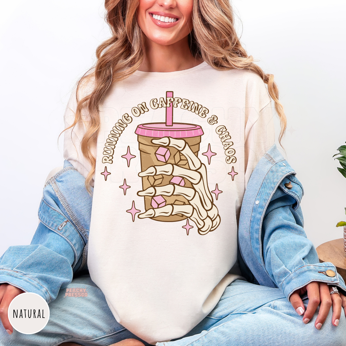 Woman wearing a natural shirt with a skeleton hand iced coffee graphic and "Running on Caffeine & Chaos" text.