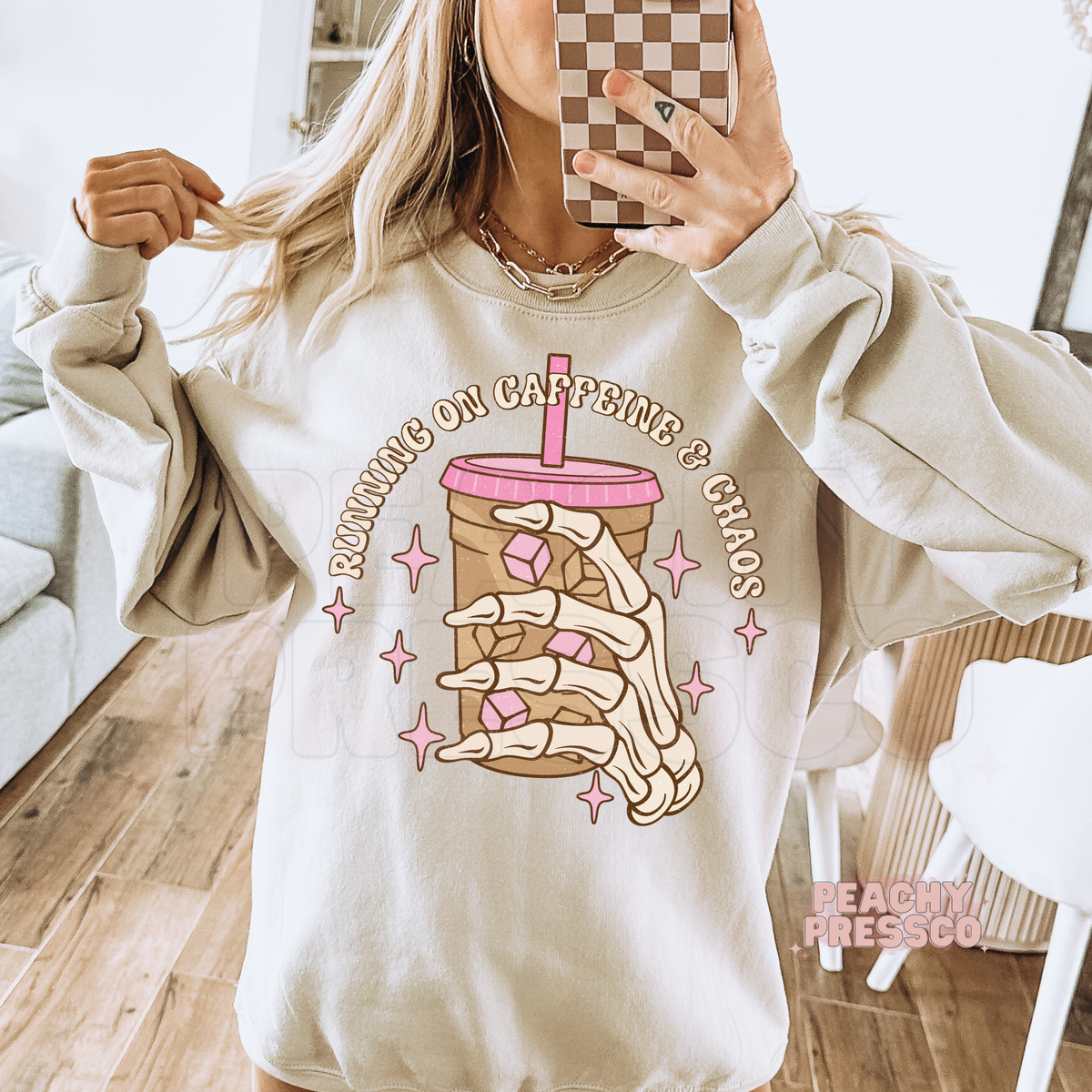 Mirror selfie of a woman wearing a sand crewneck sweatshirt featuring a skeleton hand iced coffee graphic.