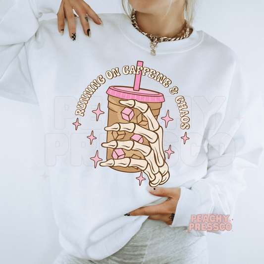 Close-up of a white sweatshirt featuring a skeleton hand iced coffee graphic with "Running on Caffeine & Chaos" lettering.