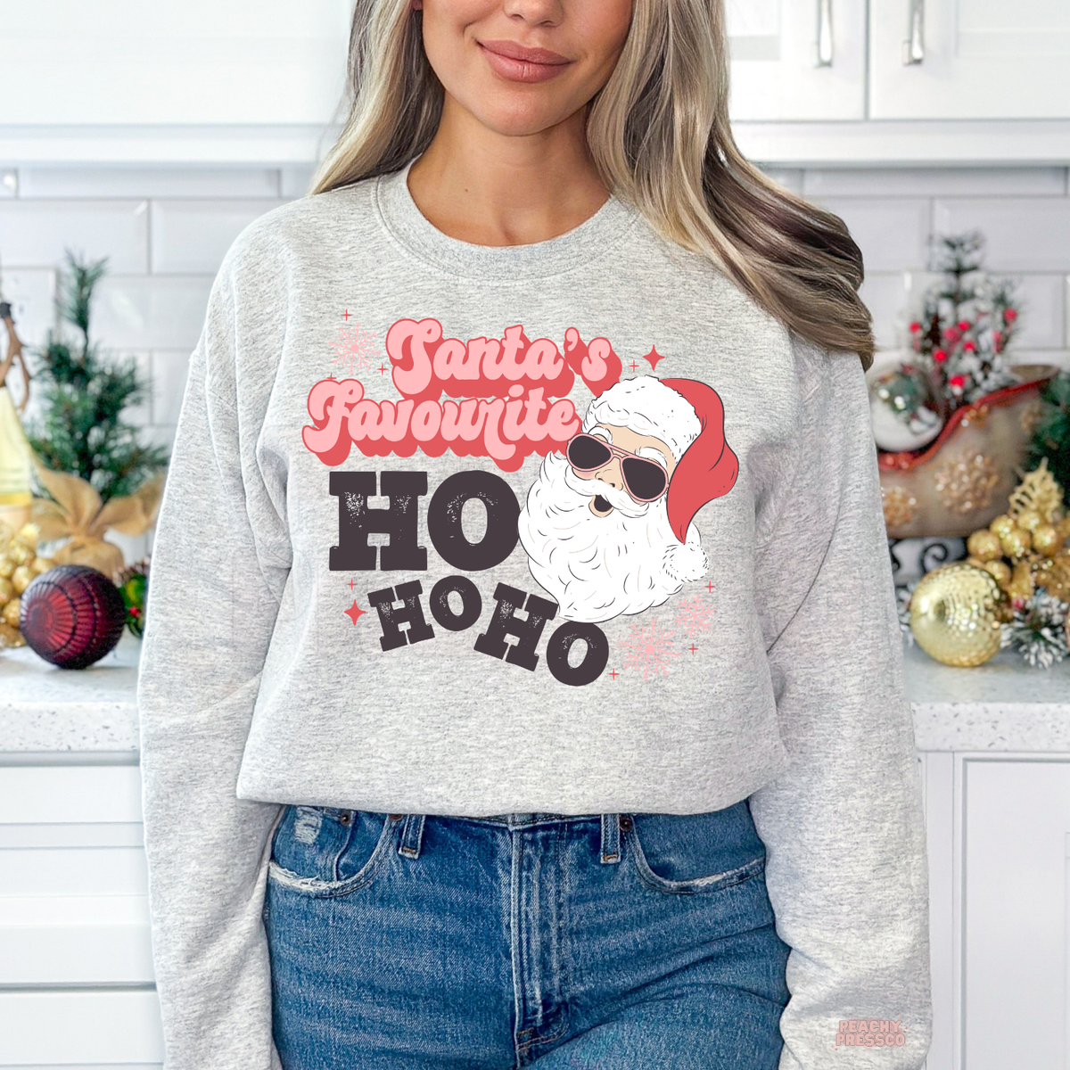 A woman wearing a ash grey crewneck sweatshirt with a festive graphic that reads 'Santa's Favourite HO HO HO' alongside an illustration of Santa Claus wearing sunglasses. She is also wearing blue jeans, standing in a decorated kitchen setting.
