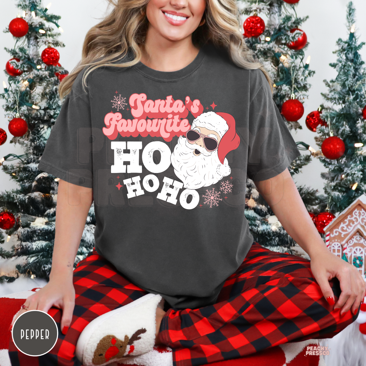 A woman wearing a comfort colors pepper graphic t-shirt that reads 'Santa's Favourite HO HO HO' with a cartoon Santa wearing sunglasses, paired with red and black plaid pajama pants, sitting in front of a decorated Christmas tree.