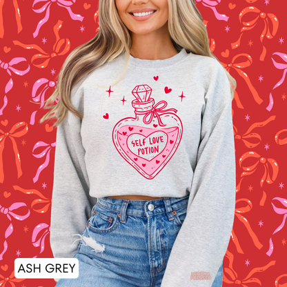 Ash grey crewneck sweatshirt with a bold Self Love Potion graphic, heart bottle artwork, and bright pink Valentine's accents.