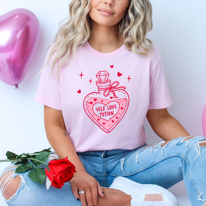 Light pink t-shirt featuring a cute Self Love Potion heart bottle graphic with pink hearts and sparkles, perfect for Valentine's Day and self-love vibes.