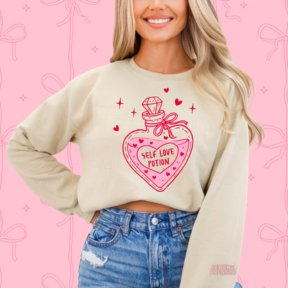 Sand crewneck sweatshirt displaying a whimsical Self Love Potion bottle design with heart details and bright pink Valentine's accents.