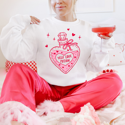 White crewneck sweatshirt featuring a fun Self Love Potion bottle graphic with pink hearts and sparkles, styled for a cozy Valentine's Day look.