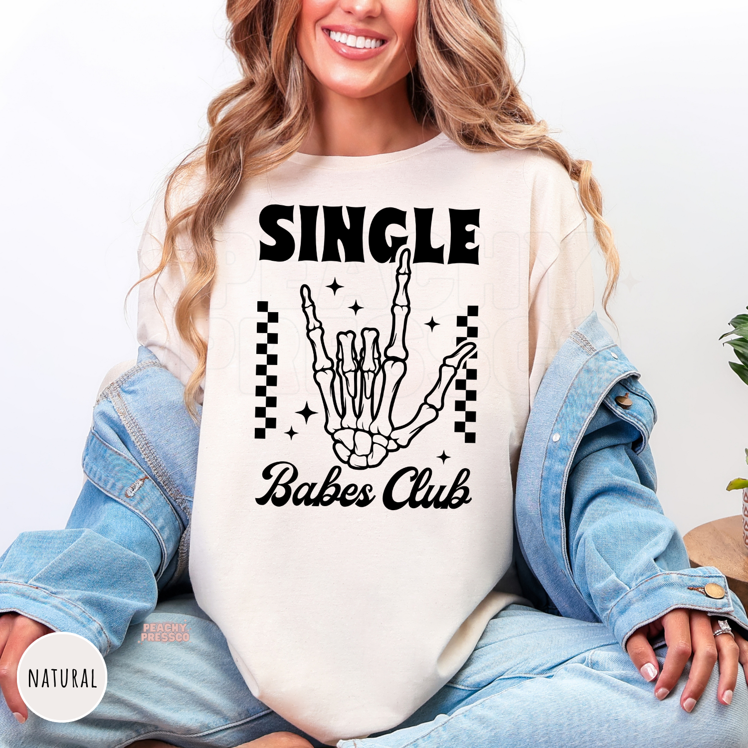 Natural shirt featuring a bold "Single Babes Club" skeleton hand graphic with retro lettering, checkerboard accents, and sparkles for Valentine's Day.