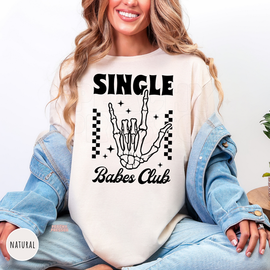 Natural shirt featuring a bold "Single Babes Club" skeleton hand graphic with retro lettering, checkerboard accents, and sparkles for Valentine's Day.