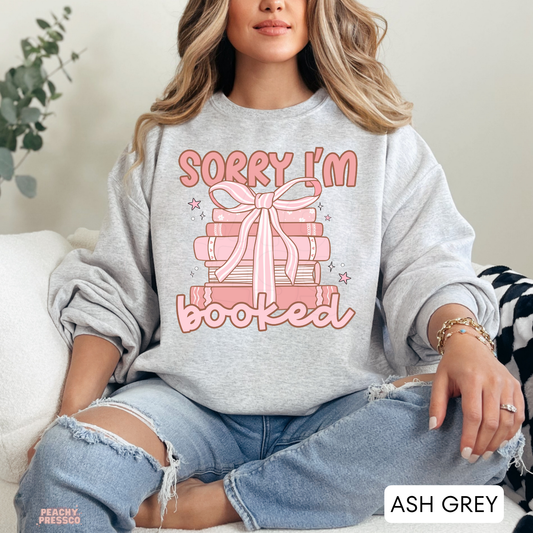 Ash Grey crewneck sweatshirt featuring a pink stack of books with a bow and "Sorry I'm Booked" design for bookworms and cozy readers.