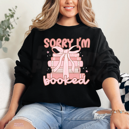 Black crewneck sweatshirt featuring a pink stack of books with a bow and "Sorry I'm Booked" graphic, a cute gift for book lovers.
