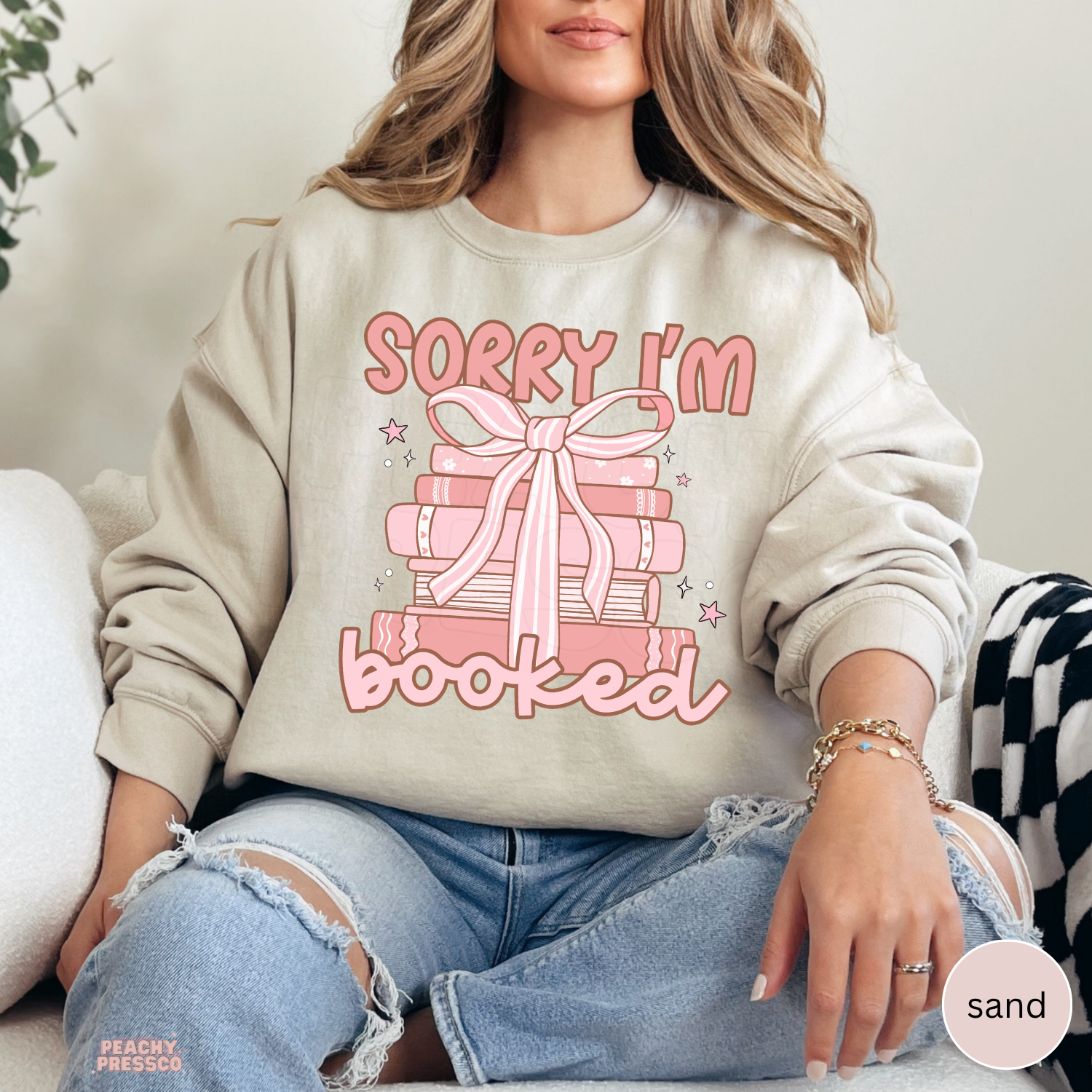 Sand crewneck sweatshirt featuring a pink stack of books with a ribbon bow and "Sorry I'm Booked" design for book lovers.