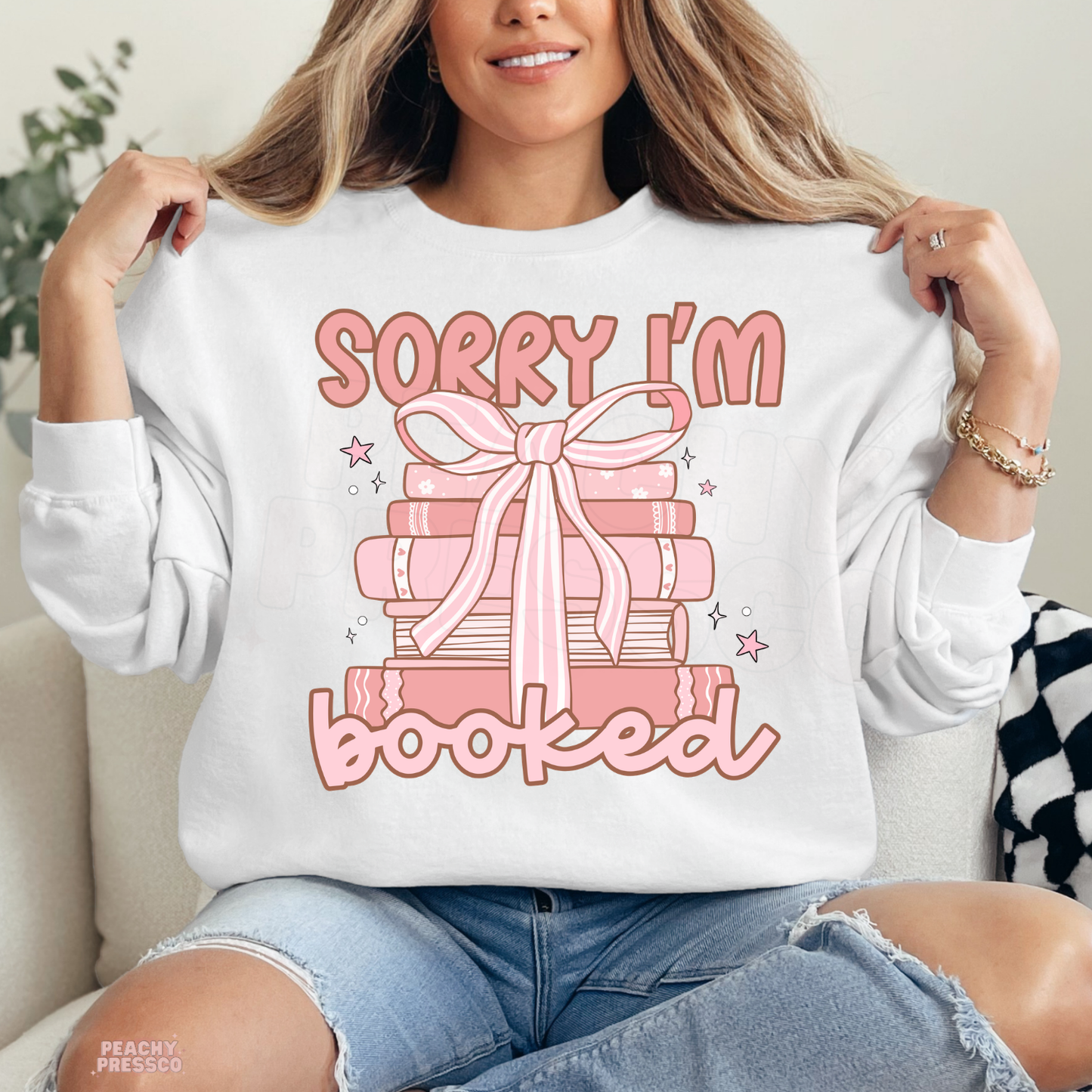 White crewneck sweatshirt with a pink book stack tied with a bow and "Sorry I'm Booked" graphic, perfect for reading lovers and book clubs.