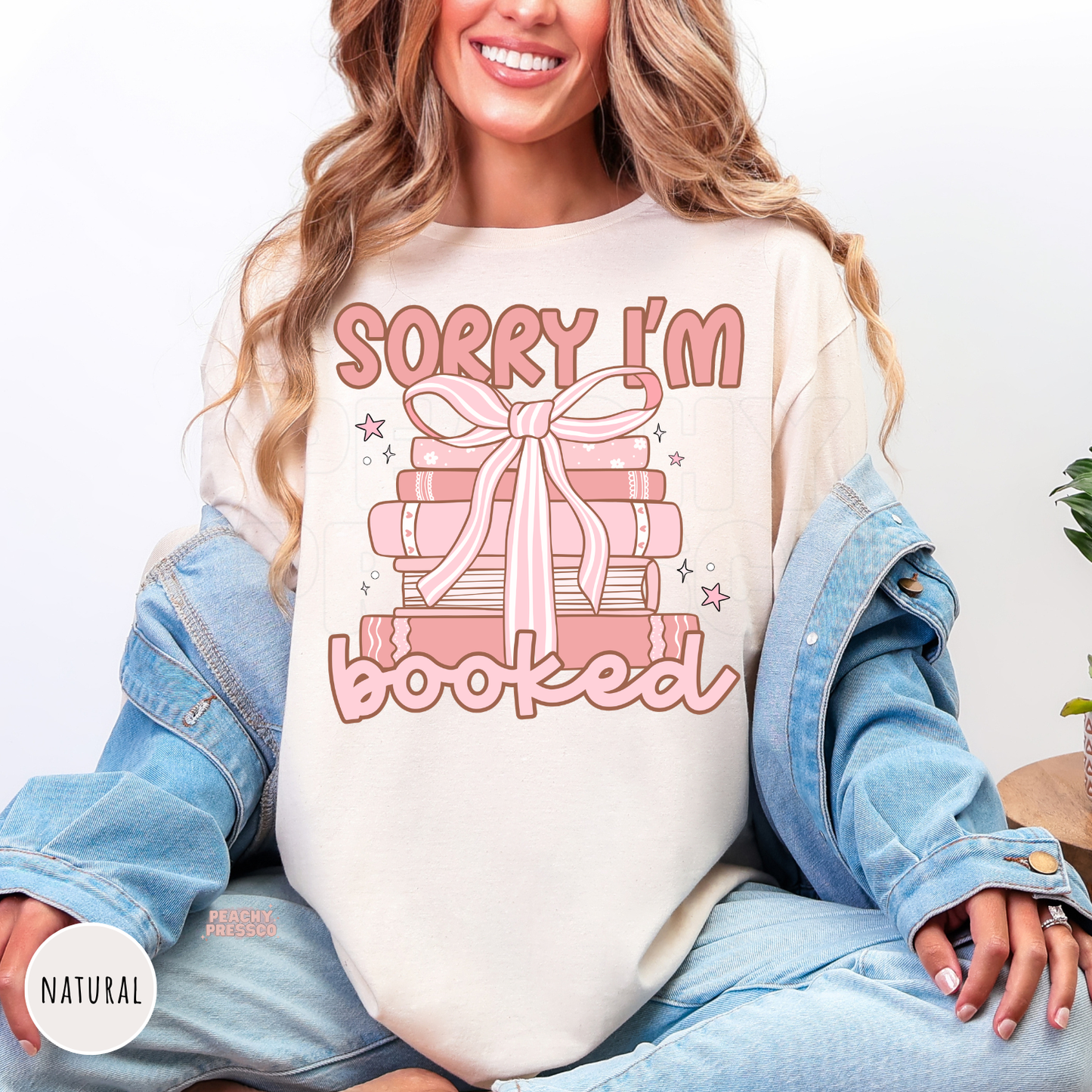Natural unisex graphic tee featuring a pink stack of books tied with a bow and the phrase "Sorry I'm Booked," perfect for book lovers and readers.