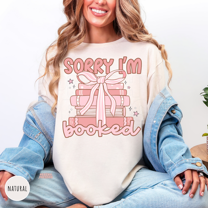Natural unisex graphic tee featuring a pink stack of books tied with a bow and the phrase "Sorry I'm Booked," perfect for book lovers and readers.