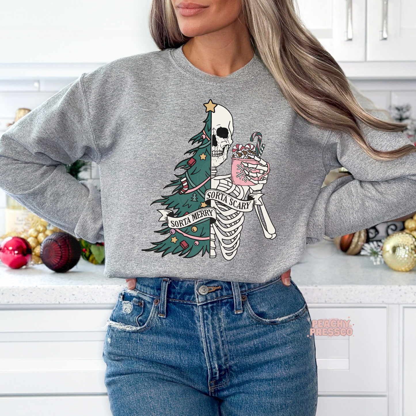 A woman wearing a sport grey crewneck sweatshirt with a festive Santa skeleton and Christmas tree graphic, paired with blue jeans in a bright kitchen setting.