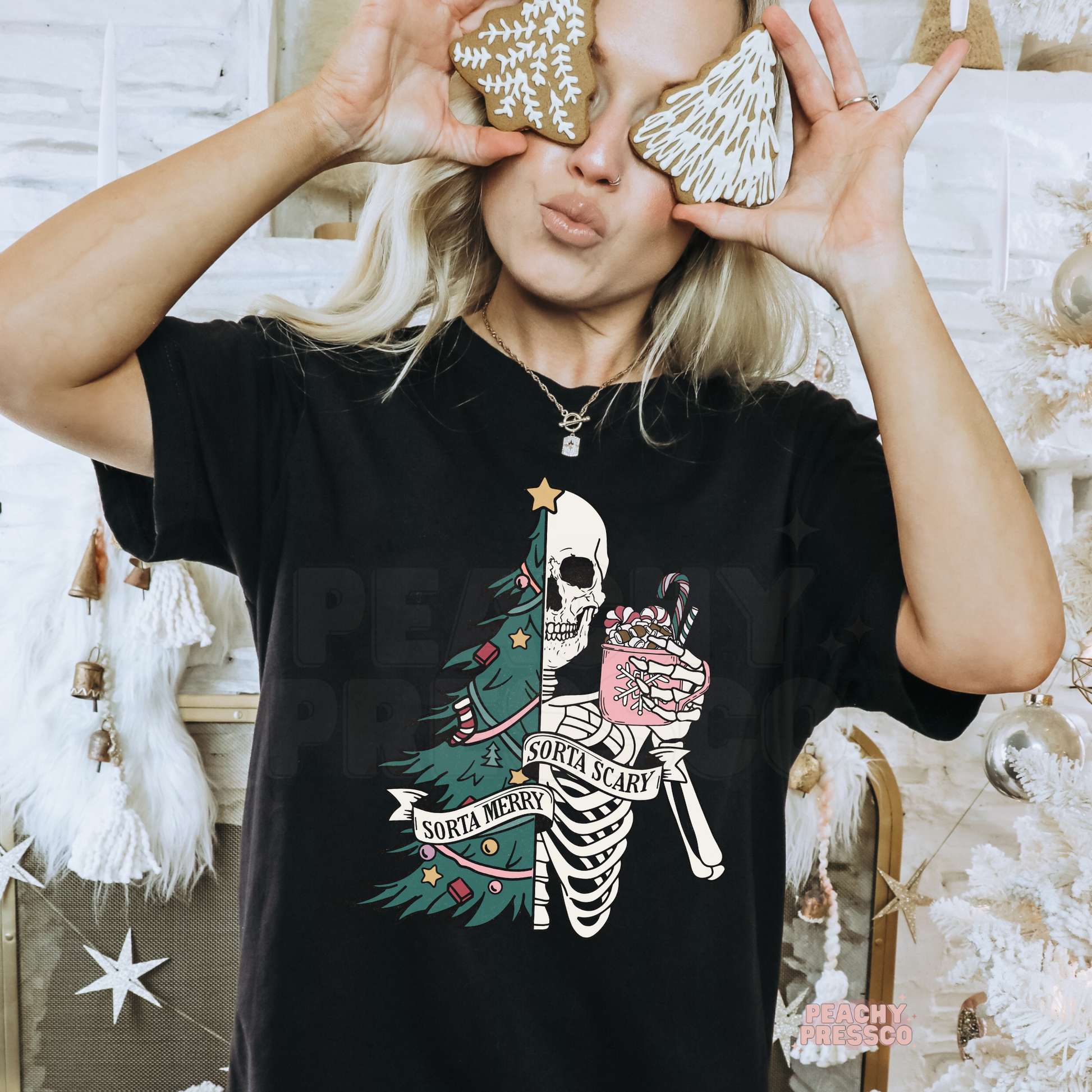 A woman wearing a black graphic t-shirt with a Christmas skeleton and tree design, holding gingerbread cookies over her eyes in a festive indoor setting.