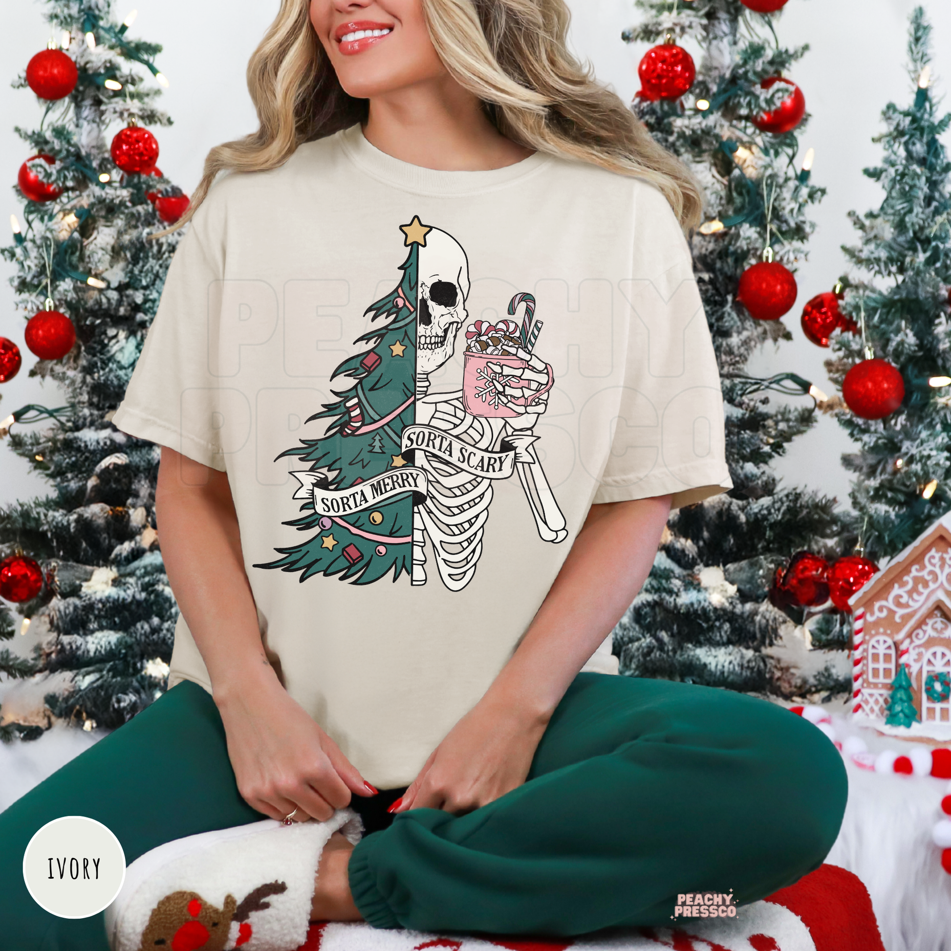A woman wearing an ivory graphic t-shirt with a Christmas skeleton and tree design, paired with green leggings, sitting in front of decorated Christmas trees.