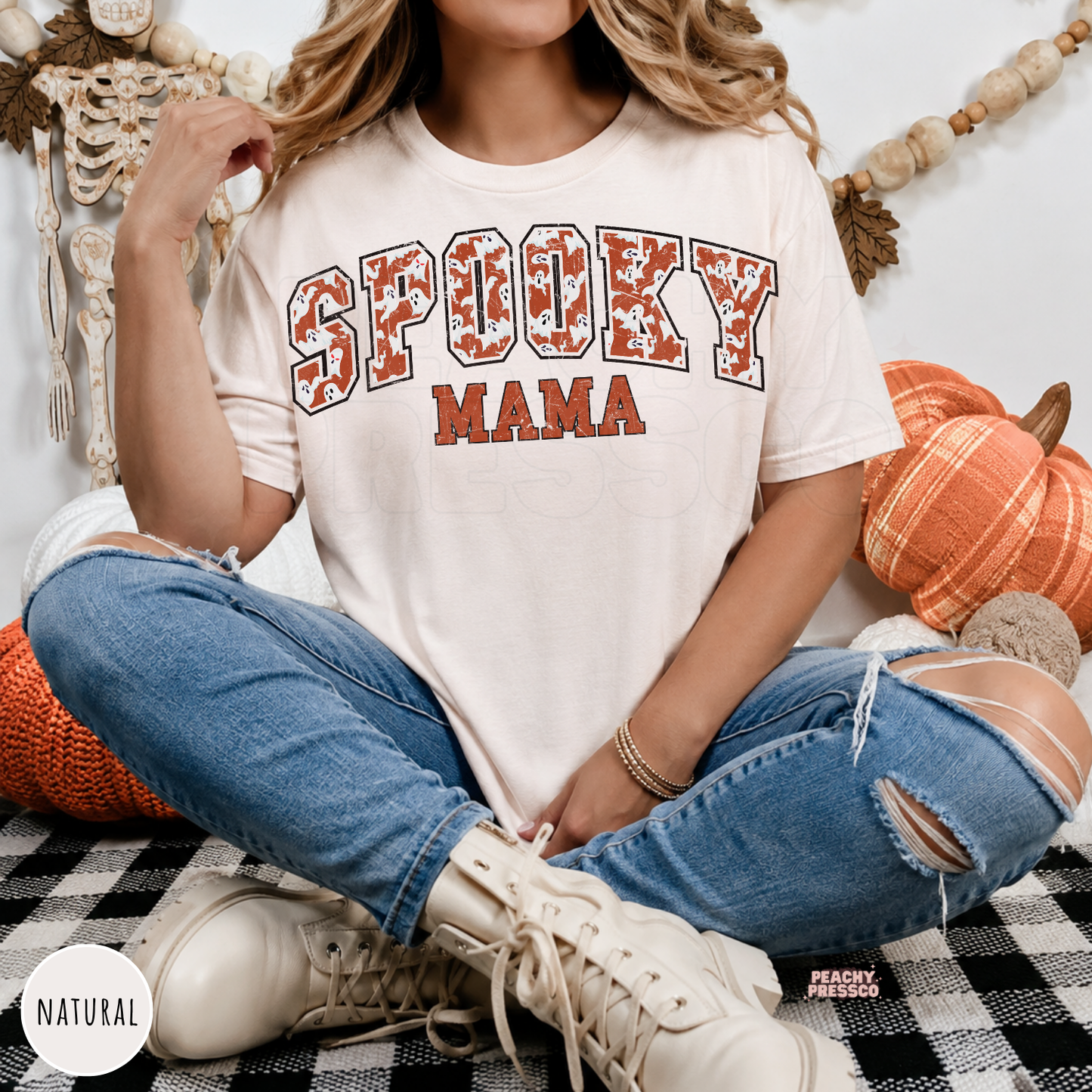 Woman wearing a natural colored Spooky Mama Halloween graphic t-shirt with distressed ghost varsity design and fall décor.