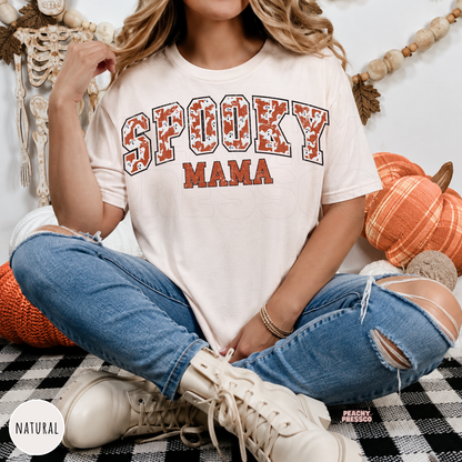 Woman wearing a natural colored Spooky Mama Halloween graphic t-shirt with distressed ghost varsity design and fall décor.
