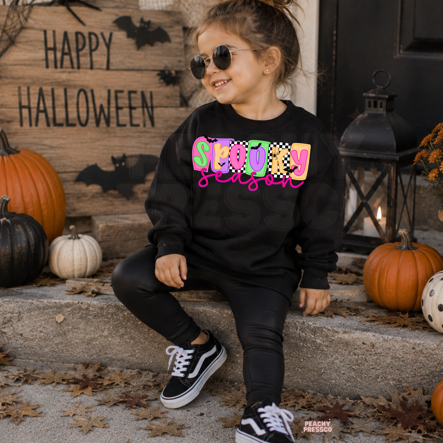 Girl wearing a black Spooky Season Halloween sweatshirt with a colorful pink, orange, green and checkerboard graphic.