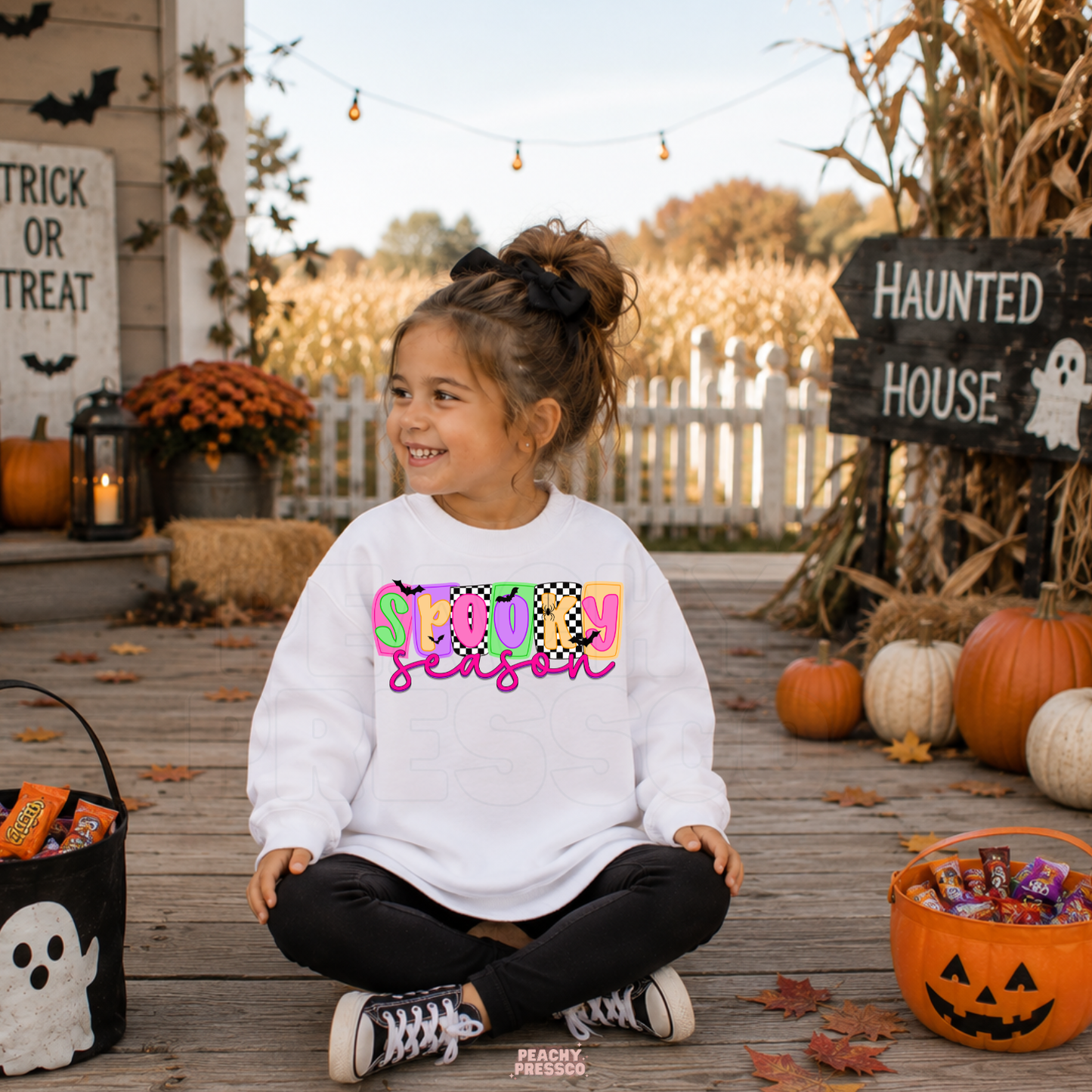 Girl wearing a white Spooky Season Halloween sweatshirt with a colorful retro graphic surrounded by pumpkins and trick-or-treat decor.