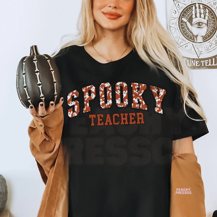 Woman wearing a black Spooky Teacher Halloween graphic t-shirt with distressed varsity ghost lettering while holding a black skeleton pumpkin.