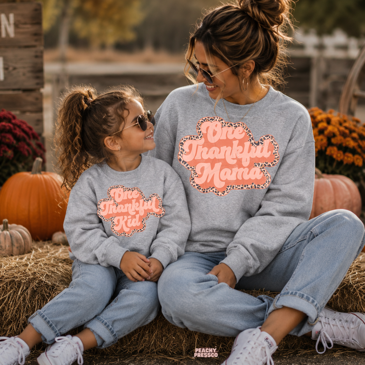 Mom and daughter smiling at each other in matching sport grey One Thankful Mama and One Thankful Kid sweatshirts at a cozy pumpkin patch.