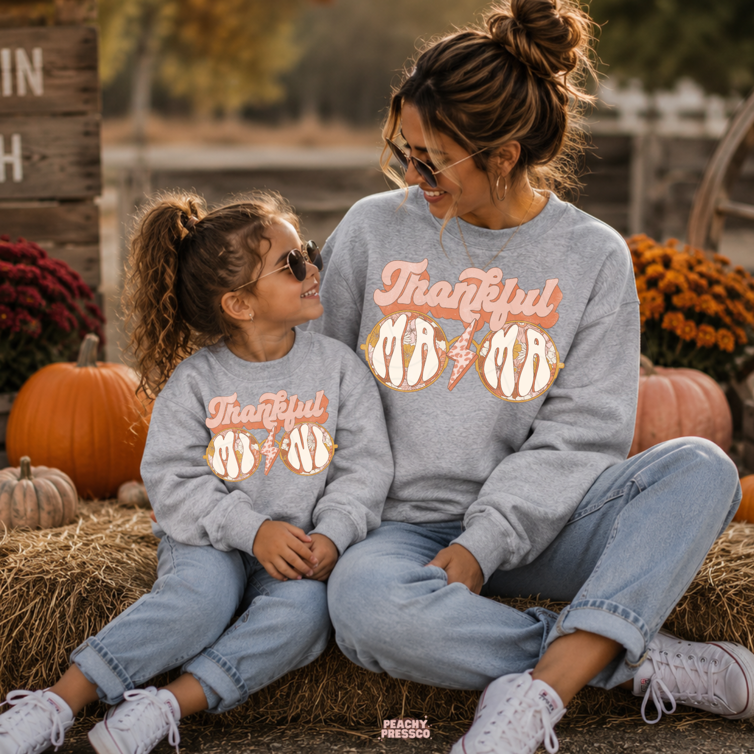 Matching sport grey Thankful Mama sweatshirts for mom and daughter sitting together at a pumpkin patch surrounded by autumn pumpkins.