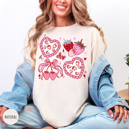 Woman wearing a natural tshirt featuring a cute coquette strawberry graphic with heart-shaped strawberry pies, bows, fresh strawberries, hearts, and pink accents.