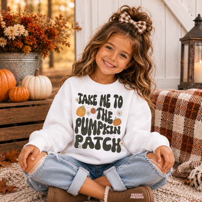 Happy toddler girl wearing a white "Take Me to the Pumpkin Patch" sweatshirt while sitting outdoors with pumpkins, fall décor, and cozy autumn decorations.