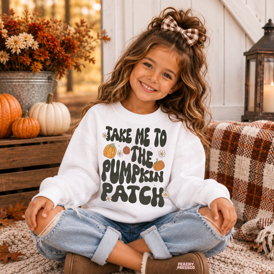 Happy toddler girl wearing a white "Take Me to the Pumpkin Patch" sweatshirt while sitting outdoors with pumpkins, fall décor, and cozy autumn decorations.