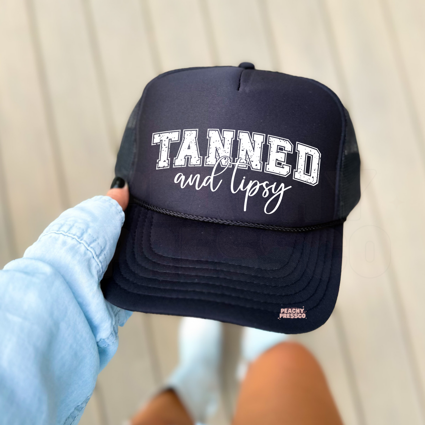 Black foam trucker hat featuring the phrase "Tanned and Tipsy" in distressed varsity lettering with elegant script. Funny summer trucker hat perfect for beach days, lake weekends, vacations, boating, bachelorette trips, and sunny getaways.