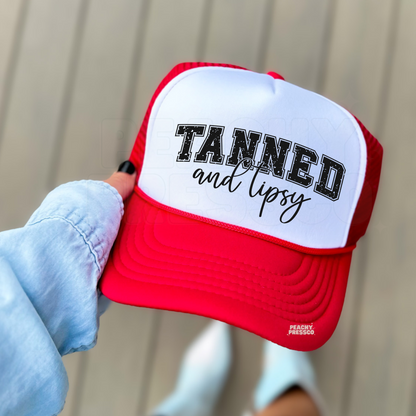 Red and white foam trucker hat featuring a distressed "Tanned and Tipsy" design in varsity lettering with script text. Fun beach trucker hat for summer vacations, lake weekends, boat days, girls' trips, and tropical getaways.