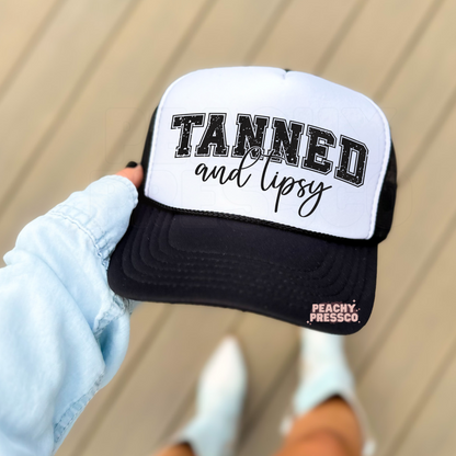 White and black foam trucker hat with a bold "Tanned and Tipsy" graphic in distressed varsity lettering and script font. Trendy summer trucker hat for beach vacations, lake life, pool parties, boating, and girls' trips.