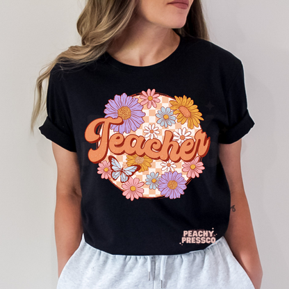 Black teacher t-shirt featuring retro floral "Teacher" lettering with colorful flowers, butterfly, and peach checkerboard background.