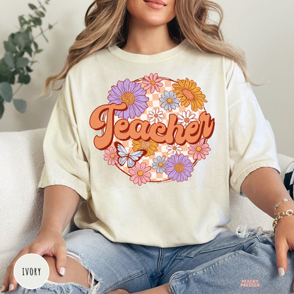 Ivory Comfort Colors teacher t-shirt featuring retro floral "Teacher" lettering with colorful flowers, butterfly, and peach checkerboard background.