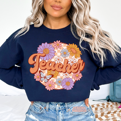 Navy teacher sweatshirt featuring a colorful retro "Teacher" floral design with pastel flowers, butterfly, and vintage checkerboard background.