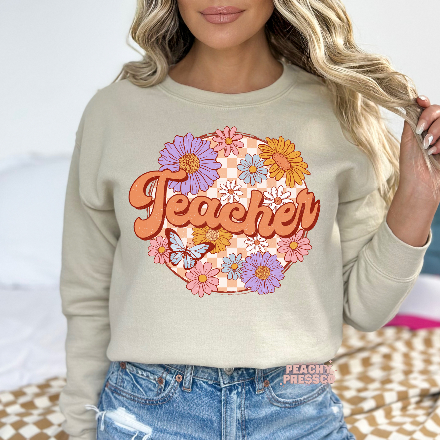 Sand teacher sweatshirt with vintage floral "Teacher" lettering, colorful flowers, butterfly, and retro peach checkerboard design.