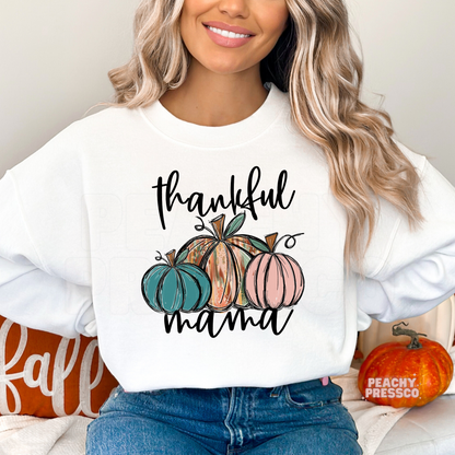 Woman wearing a white crewneck sweatshirt with a "Thankful Mama" pumpkin graphic featuring colorful pumpkins, ideal for Thanksgiving gatherings and fall season.