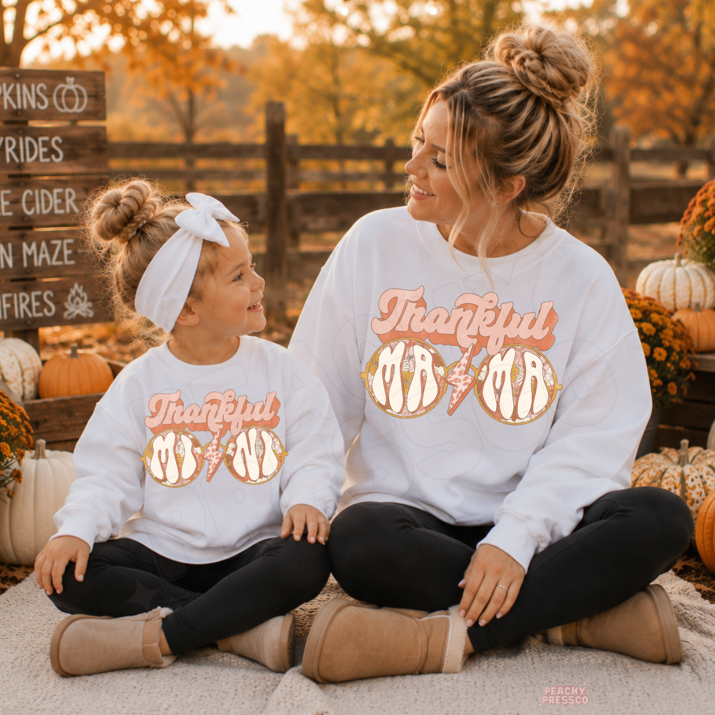 Mom and daughter wearing matching white Thankful Mama sweatshirts while smiling at each other during a cozy pumpkin patch outing.