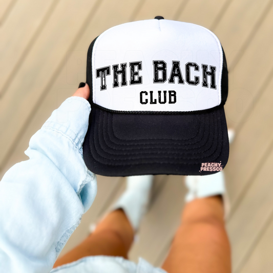 Woman holding a white and black trucker hat with "The Bach Club" distressed varsity print, trendy bachelorette party accessory for bridal weekends.