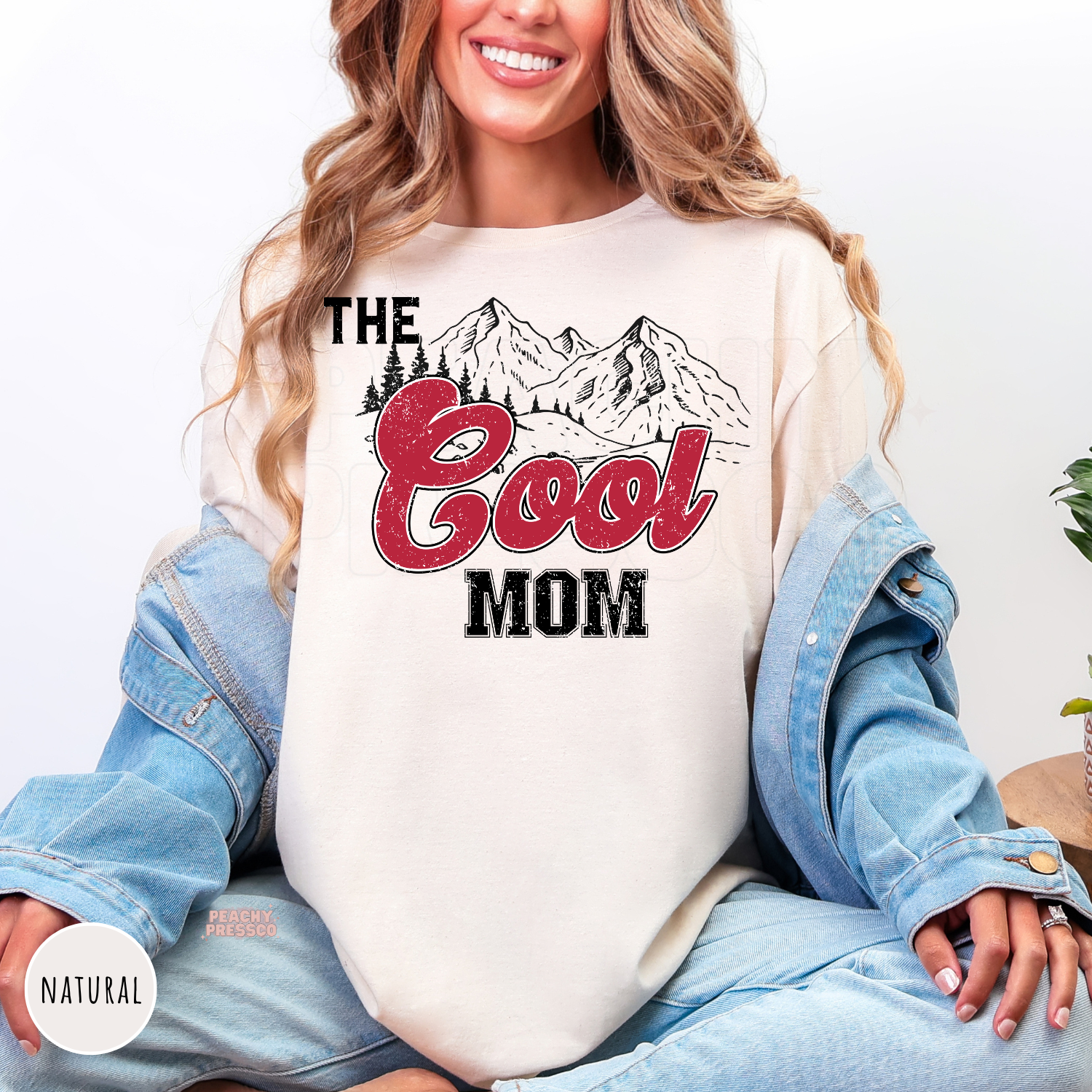 Natural graphic t-shirt featuring a vintage "The Cool Mom" mountain design with distressed red lettering. Cute western-inspired mom shirt and thoughtful Mother's Day gift.