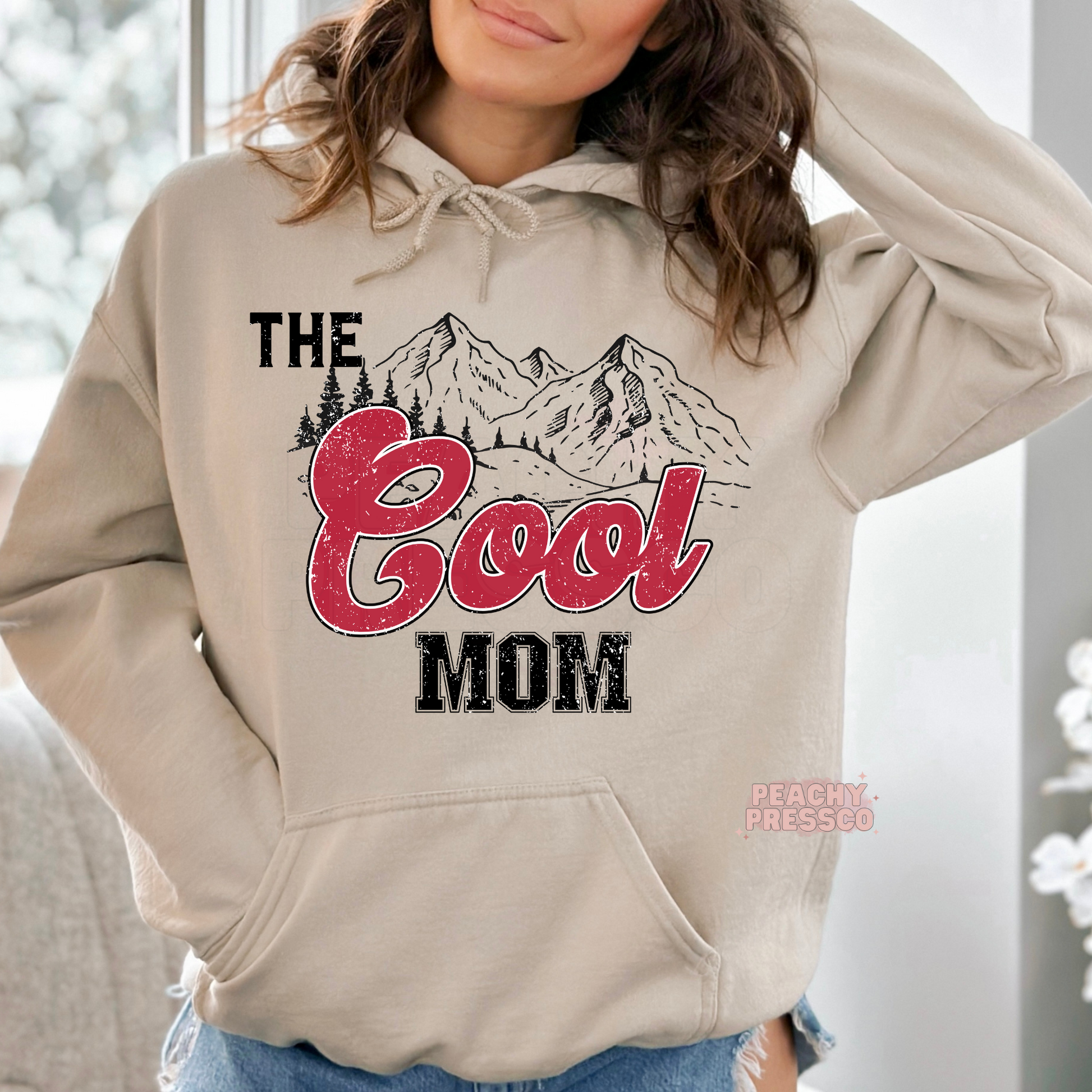 Sand hoodie featuring a retro "The Cool Mom" mountain graphic with distressed red lettering. Cozy western-inspired hoodie for stylish moms.