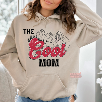 Sand hoodie featuring a retro "The Cool Mom" mountain graphic with distressed red lettering. Cozy western-inspired hoodie for stylish moms.