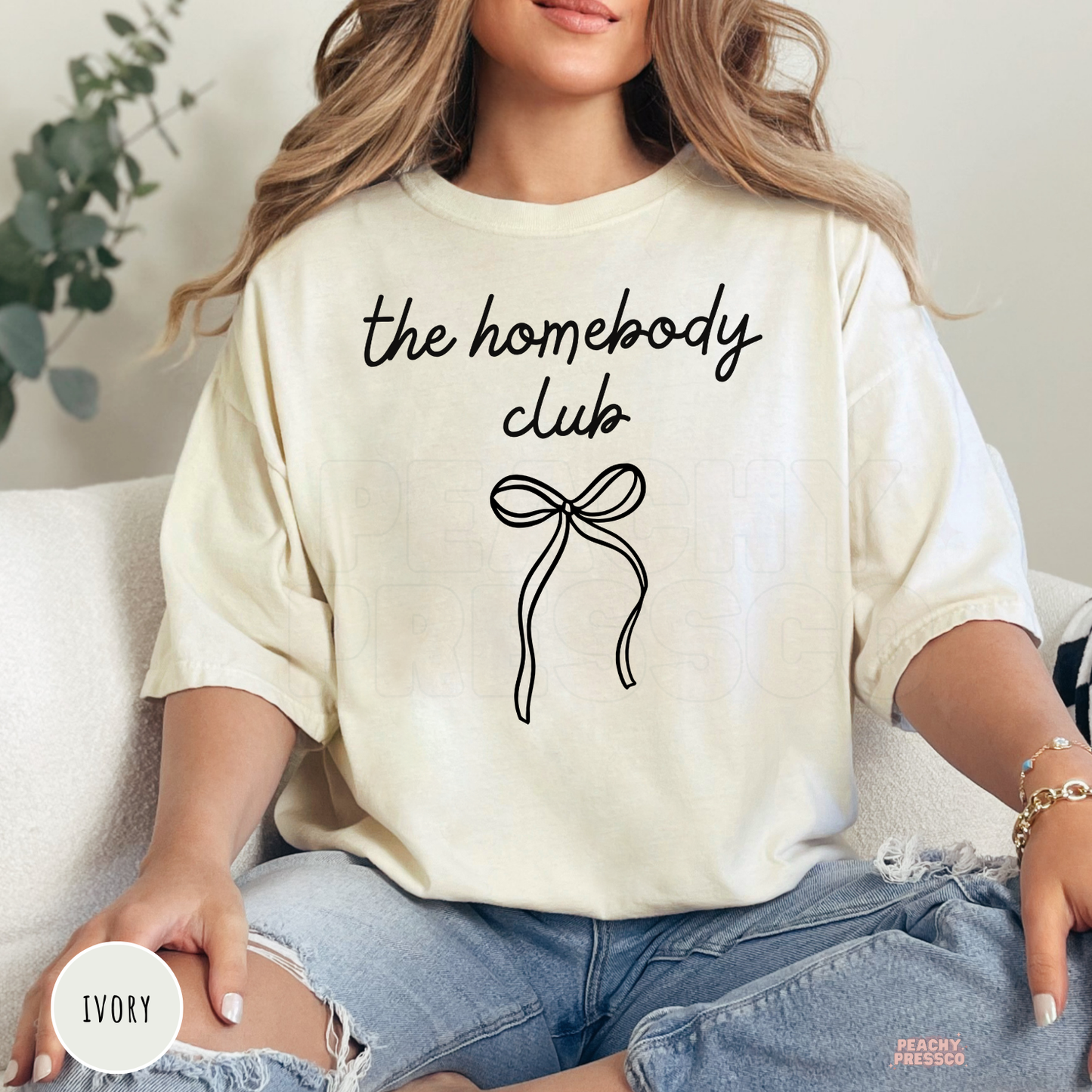 Woman wearing an ivory graphic t-shirt with "The Homebody Club" text and a minimalist black bow illustration.