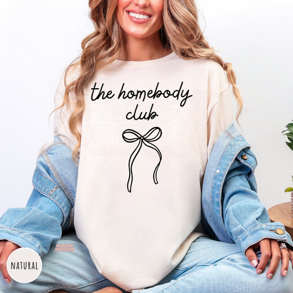Woman wearing a natural shirt featuring a minimalist "The Homebody Club" graphic with a simple coquette bow design.