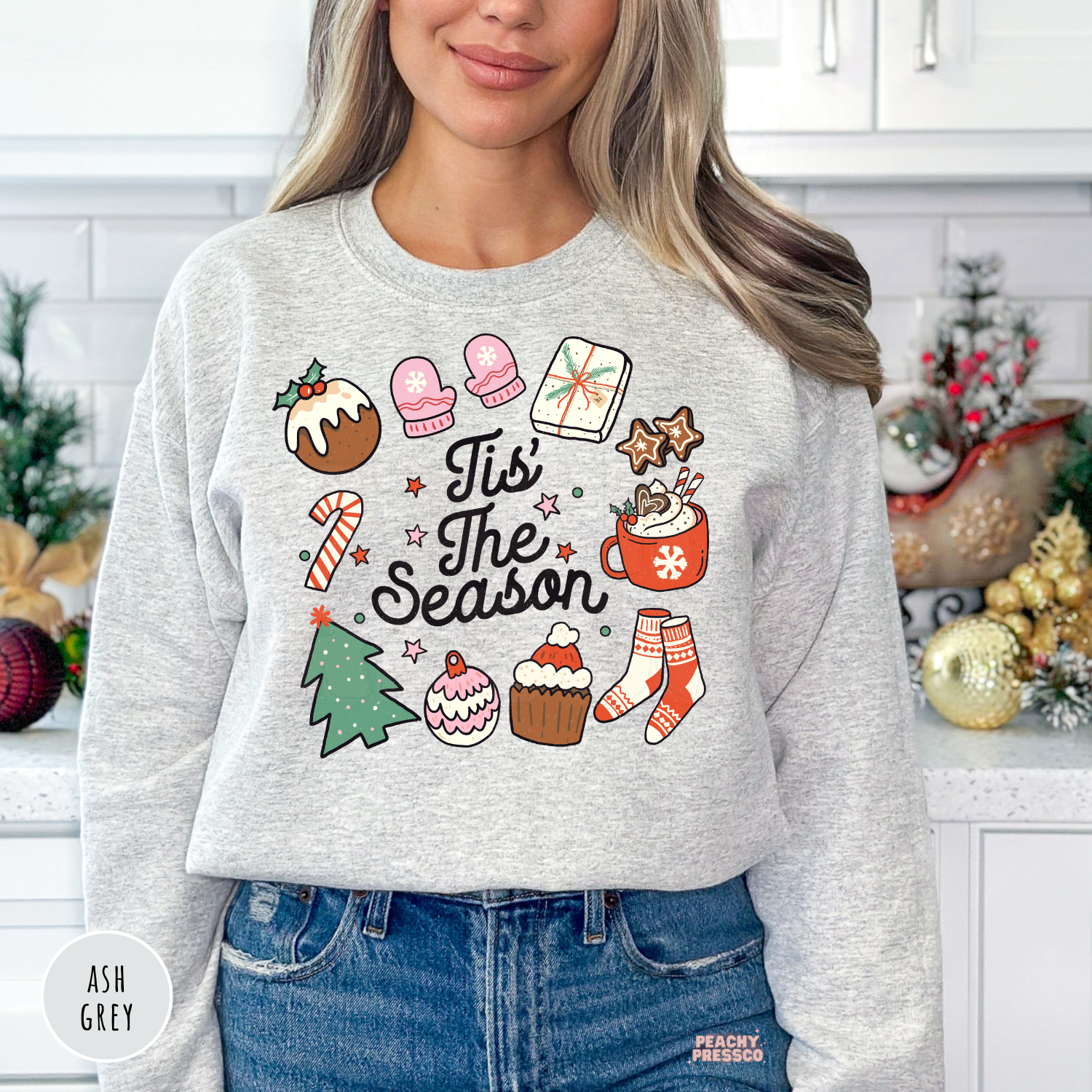 A woman wearing a ash gray long-sleeve crewneck sweatshirt with a colorful Christmas-themed graphic that reads 'Tis The Season' surrounded by festive illustrations. She is wearing blue jeans and standing in a decorated kitchen setting.